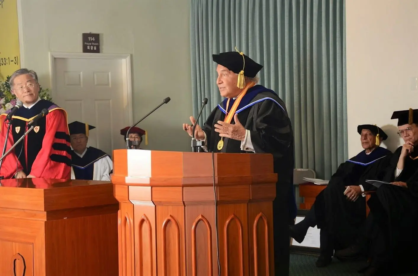 A man in graduation robes is speaking at a podium.